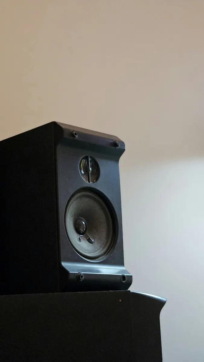 Black bookshelf loudspeaker on a dark stand against a pale wall, seen from below