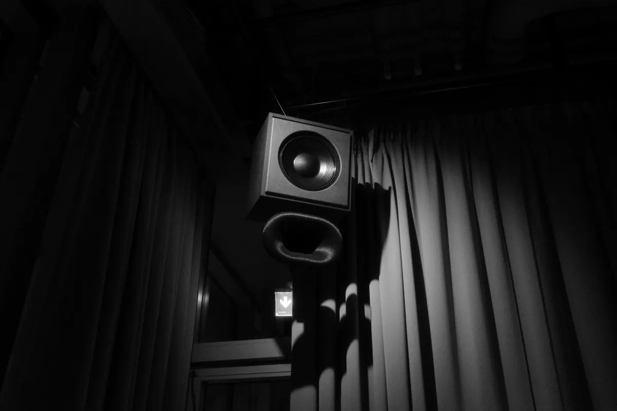 Black-and-white photo of a suspended loudspeaker with a port tube hanging in front of a heavy stage curtain